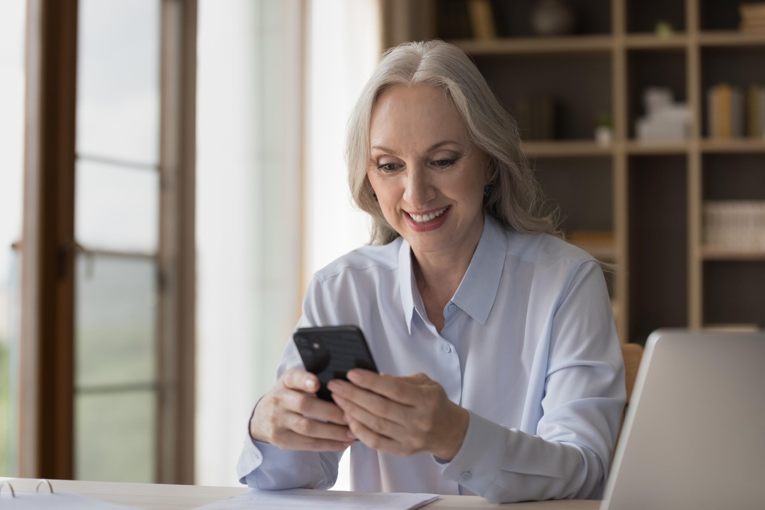 Smilingmiddle agedbusinesswomanusingsmartphoneseatedatworkplacedistractedfrom